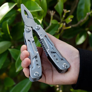 Silver Adventure Buddy multi-tool held open in one hand, pliers up, in front of green leaves