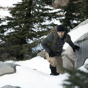 Hiker in a gray jacket and backpack climbing over snowy rocks