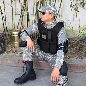 Person in gray digital camo wearing a black MOLLE vest with a flag patch, knee and elbow pads