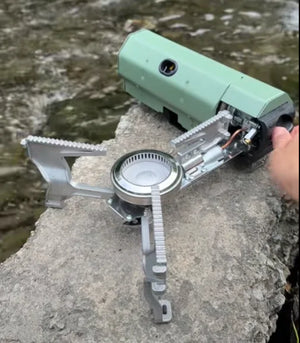 Green AdventureChef stove open on a rock by a river, a hand on the control knob