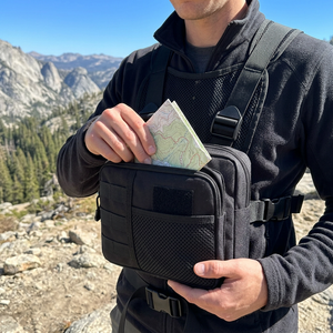 Hiker wearing a black chest rig bag with mesh harness, pulling a trail map from the front pocket