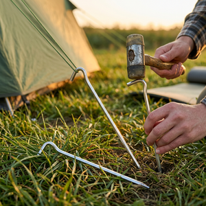 Silver GripHook aluminum hook stakes at a campsite, one holding a tent guy line while a camper taps another in with a mallet