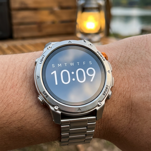 SummitNav GPS smartwatch with a silver steel link bracelet on a wrist at a lakeside campsite
