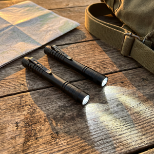 Two PenBeam black pen lights, both switched on, on a wooden camp table beside a map and pack strap