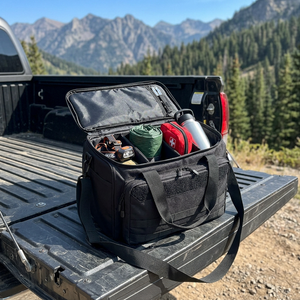 Black padded gear bag open on a truck tailgate, packed with camping gear at a mountain trailhead