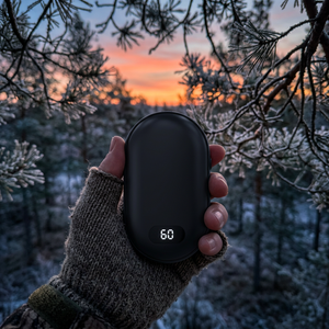 Black rechargeable hand warmer showing 60 in a gloved hand among frosty pine branches at sunset