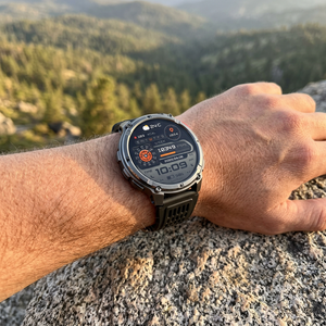 TrailCore smartwatch with all-black band on a hiker's wrist resting on a granite boulder at a mountain overlook