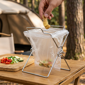 Stainless steel wire trash bag holder with a clear bag on a camp table, food scraps going in