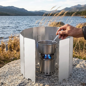 Silver WindWall folding windshield wrapped around a camp stove on a granite rock by a lake, a hand stirring the pot