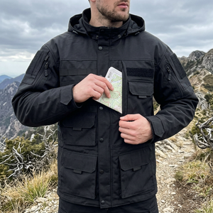 Man on a mountain trail in a black hooded tactical jacket, tucking a map into a chest pocket