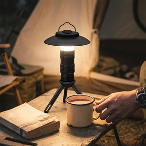 Matte Black Bright Buddy mini lantern on its tripod glowing warm beside a mug of cocoa in a tent