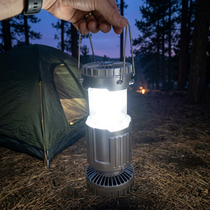 Hand carrying the silver TrailBright solar lantern, pulled up and glowing, past a tent at dusk