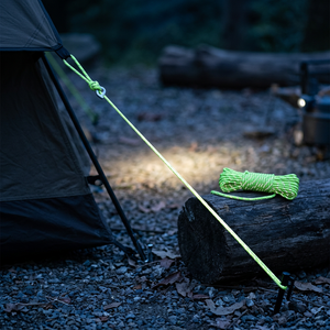 Lime green NightTrace reflective cord used as a tent guy line at dusk, with a coil of cord on a log