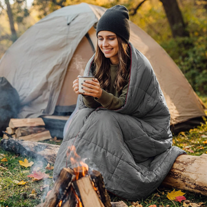 Camper wrapped in the Wolf Grey Adventure Cozy Puff wearable blanket by a campfire with a warm mug