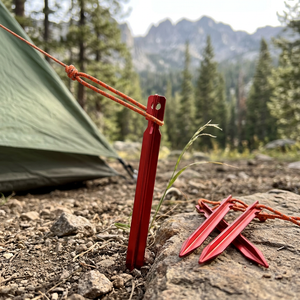 Red TriPeg Y-beam tent stake driven into rocky ground holding a tent guy line, two more stakes on a rock, mountains behind